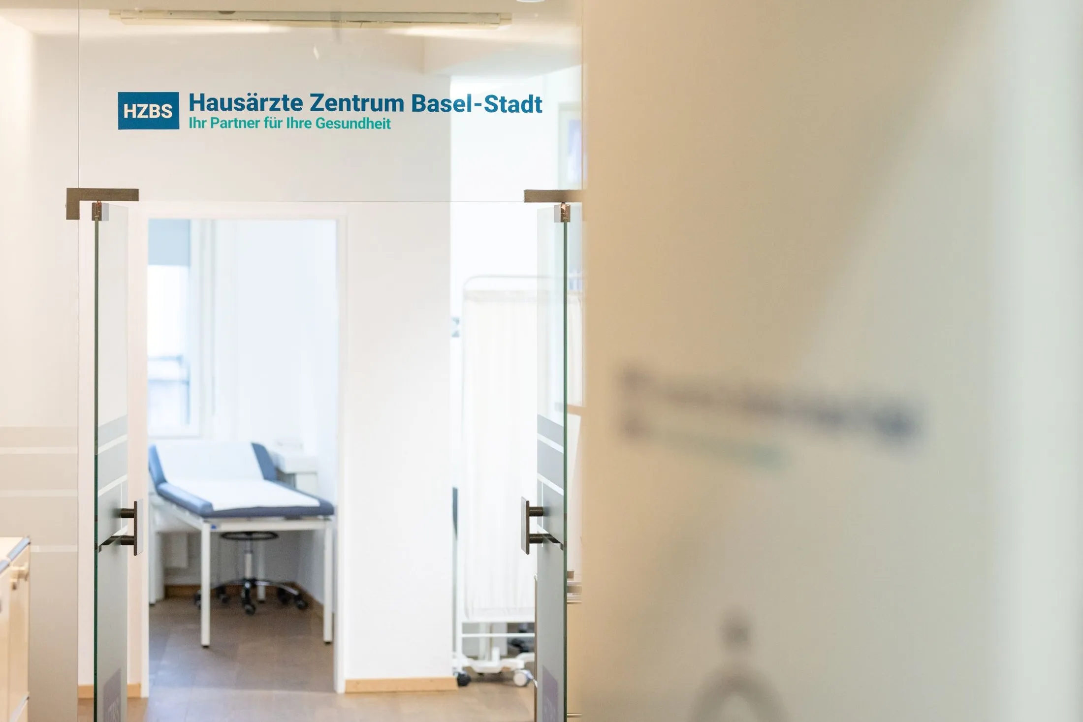 Glastür mit dem Logo des Hausärzte Zentrum Basel-Stadt, dahinter ein Sprechzimmer